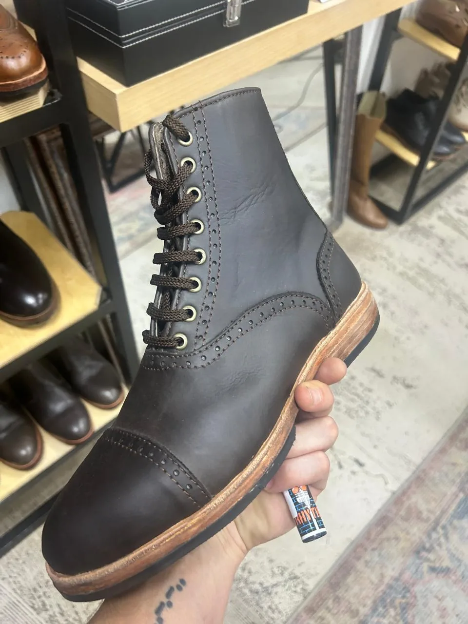 A hand holding a dark brown brogue boot with a natural sole edge, in the Atelier