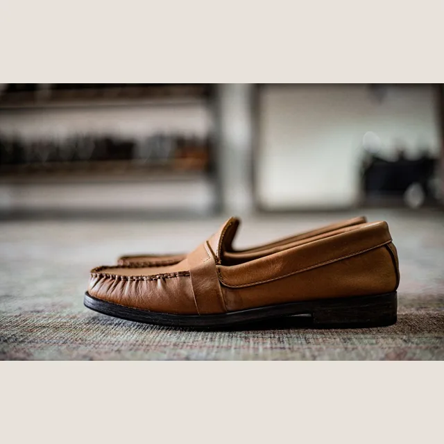 A tan loafer on a rug, shelves of pairs behind