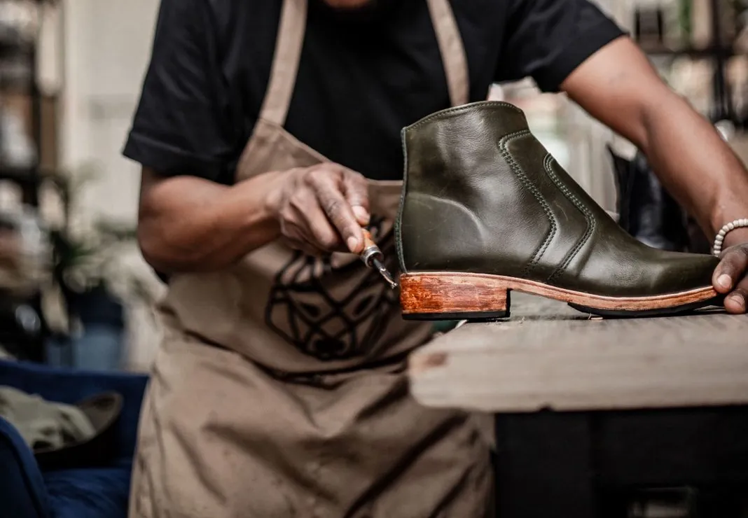 Hands at the bench working the welt of a green ankle boot with an awl