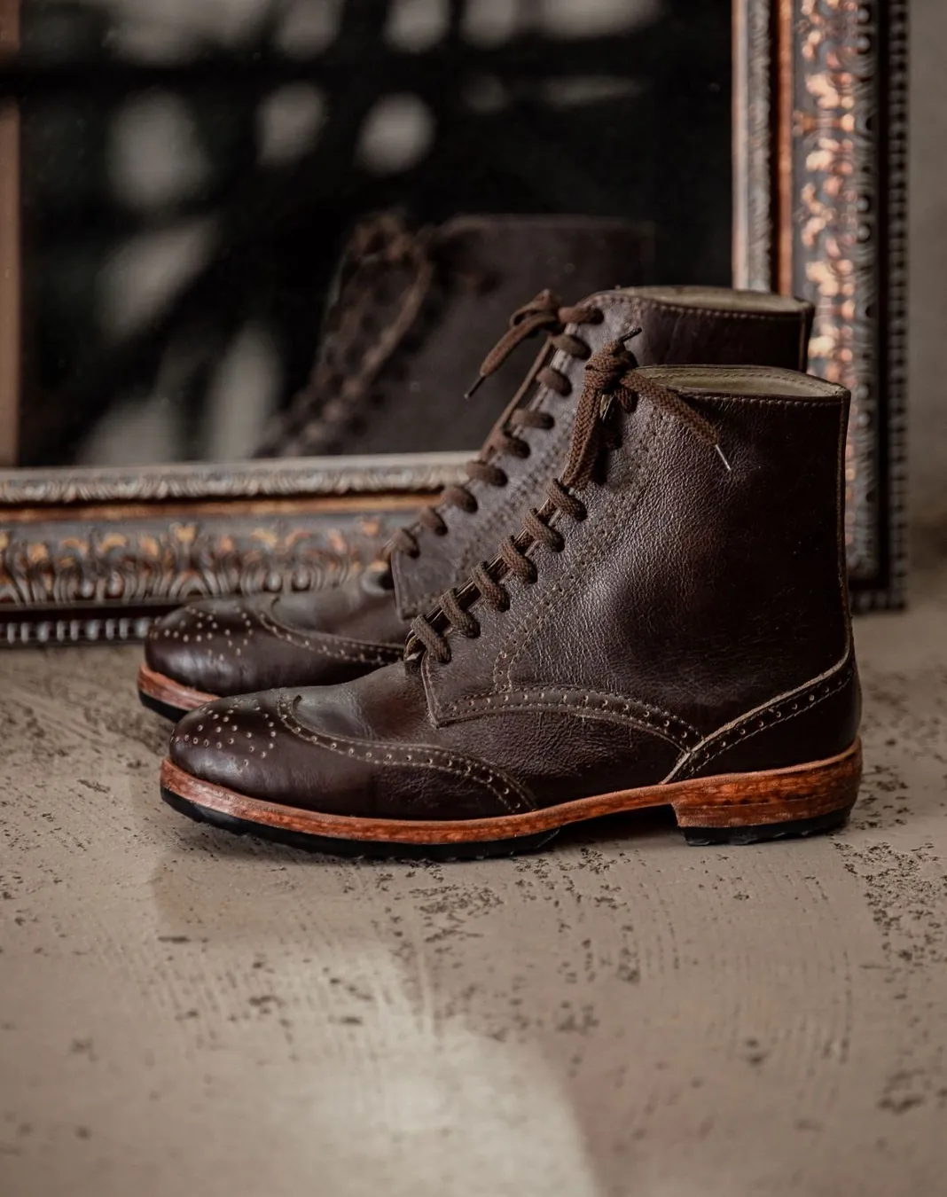 A worn pair of dark brown brogue boots with natural sole edges, standing before an old mirror